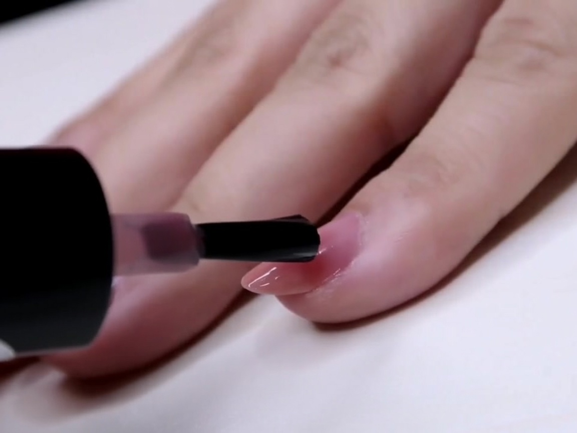 Ein Pinsel trägt eine Schicht transparenten rosa Lacks auf die äußere Hälfte eines Fingernagels auf.