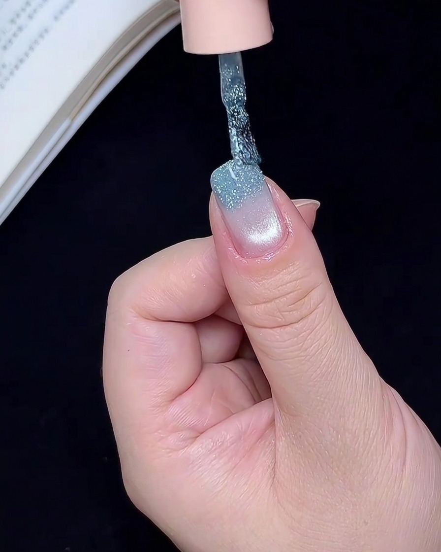 Ein Nailart-Pinsel trägt schimmerndes blaues Glitzergel auf die freie Kante eines Nagels auf.