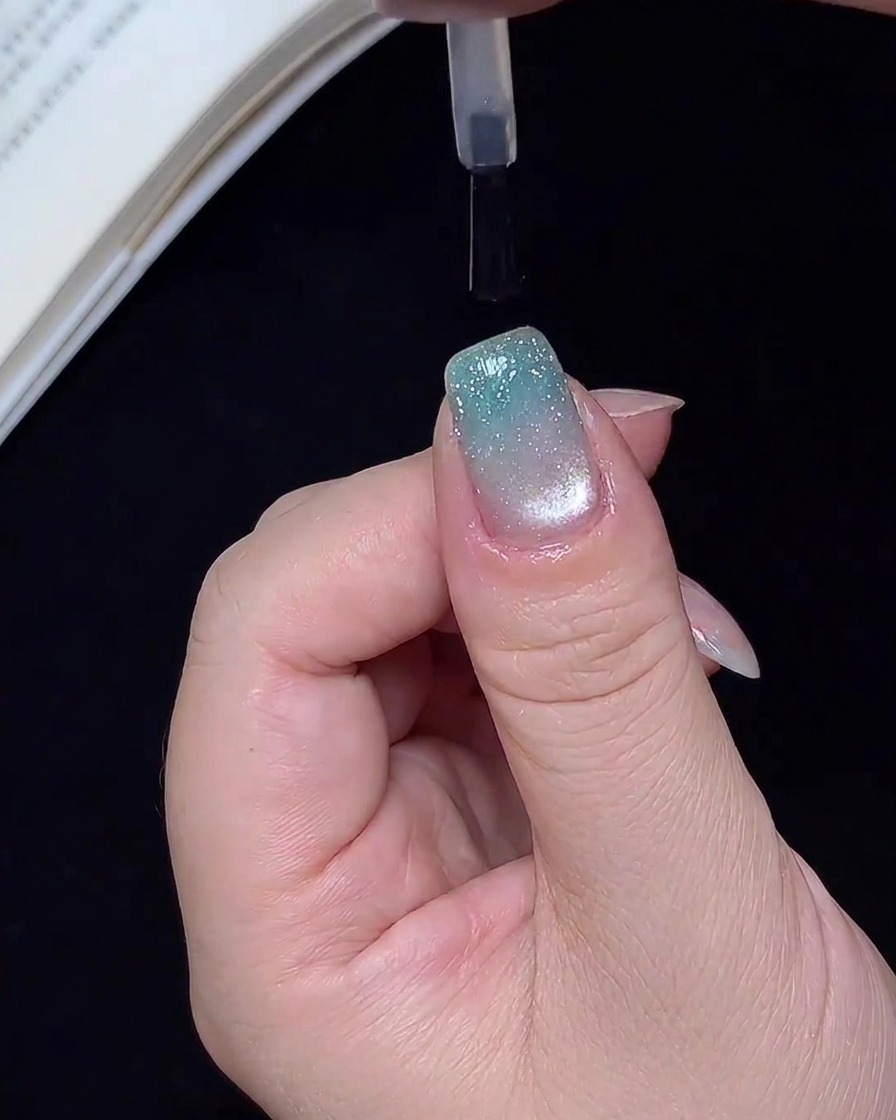 Ein Pinsel trägt einen klaren, glänzenden Überlack auf einen fertigen blauen Glitzer-Ombré-Nagel auf.