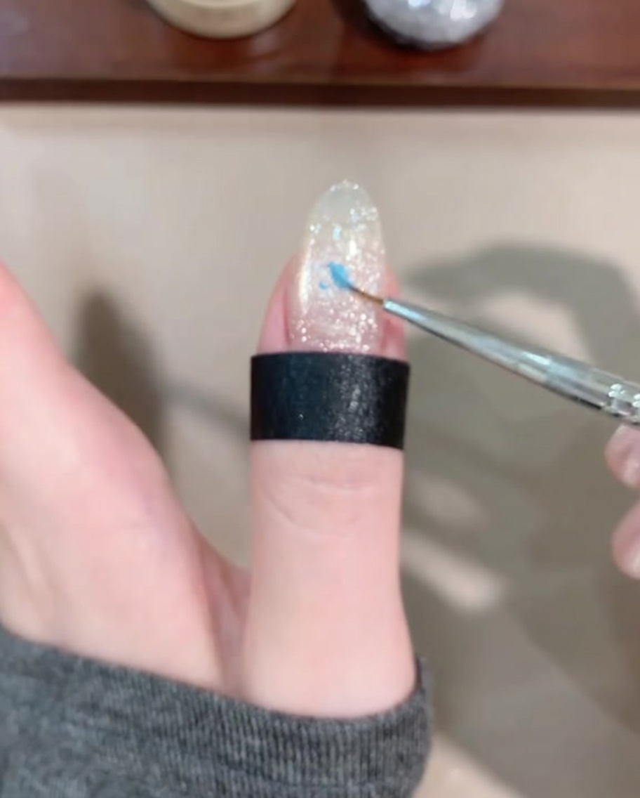Ein feiner Liner-Pinsel trägt einen kleinen Tropfen blaues Gel auf die Mitte des Nagels auf.