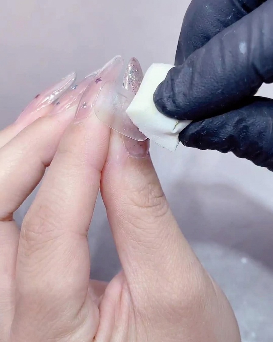 Eine behandschuhte Hand drückt einen kleinen weißen Make-up-Schwamm auf die Oberfläche eines gelbeschichteten Nagels.