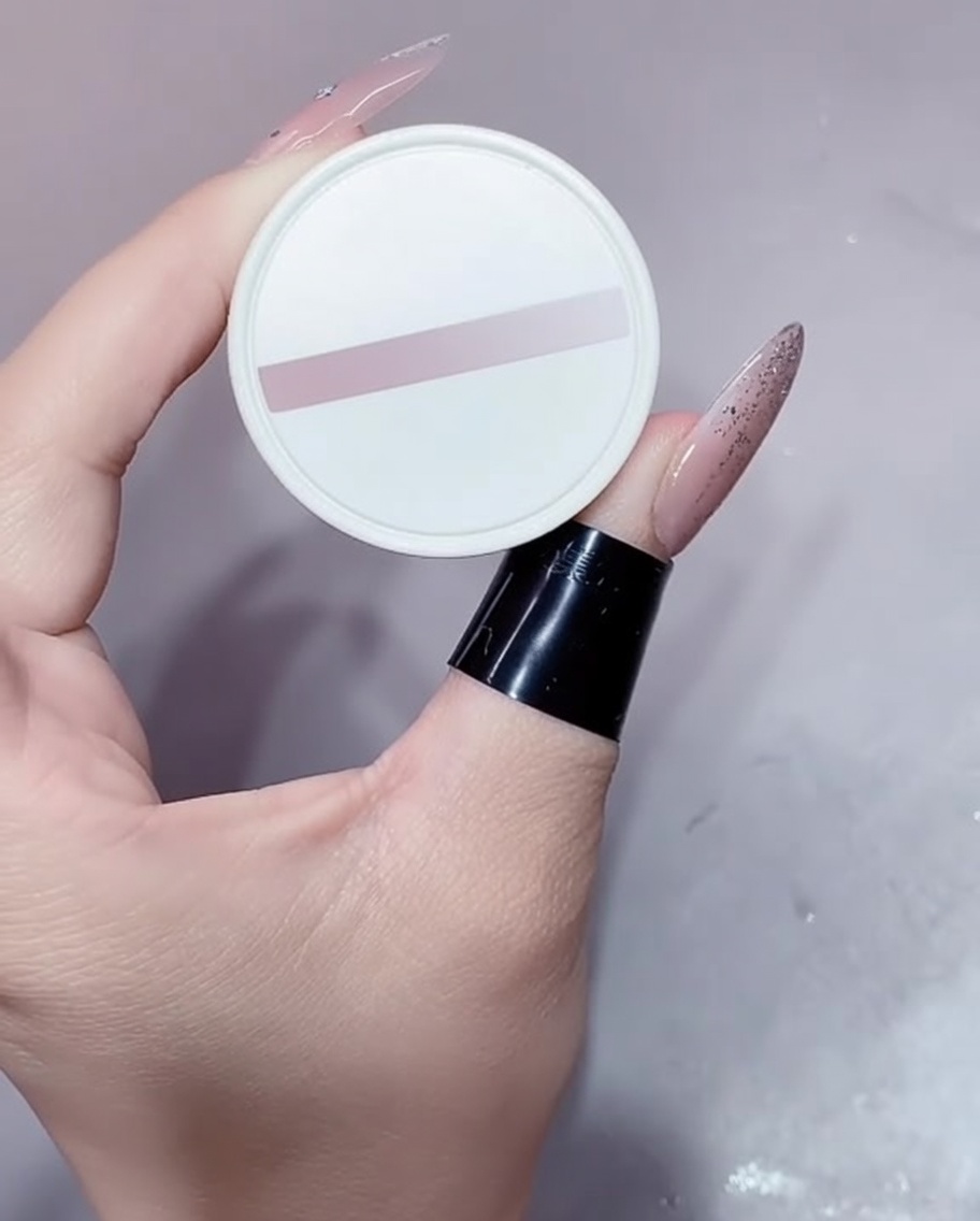 Ein kleiner weißer Tiegel mit Nude-Gel, gehalten von einer manikürten Hand.