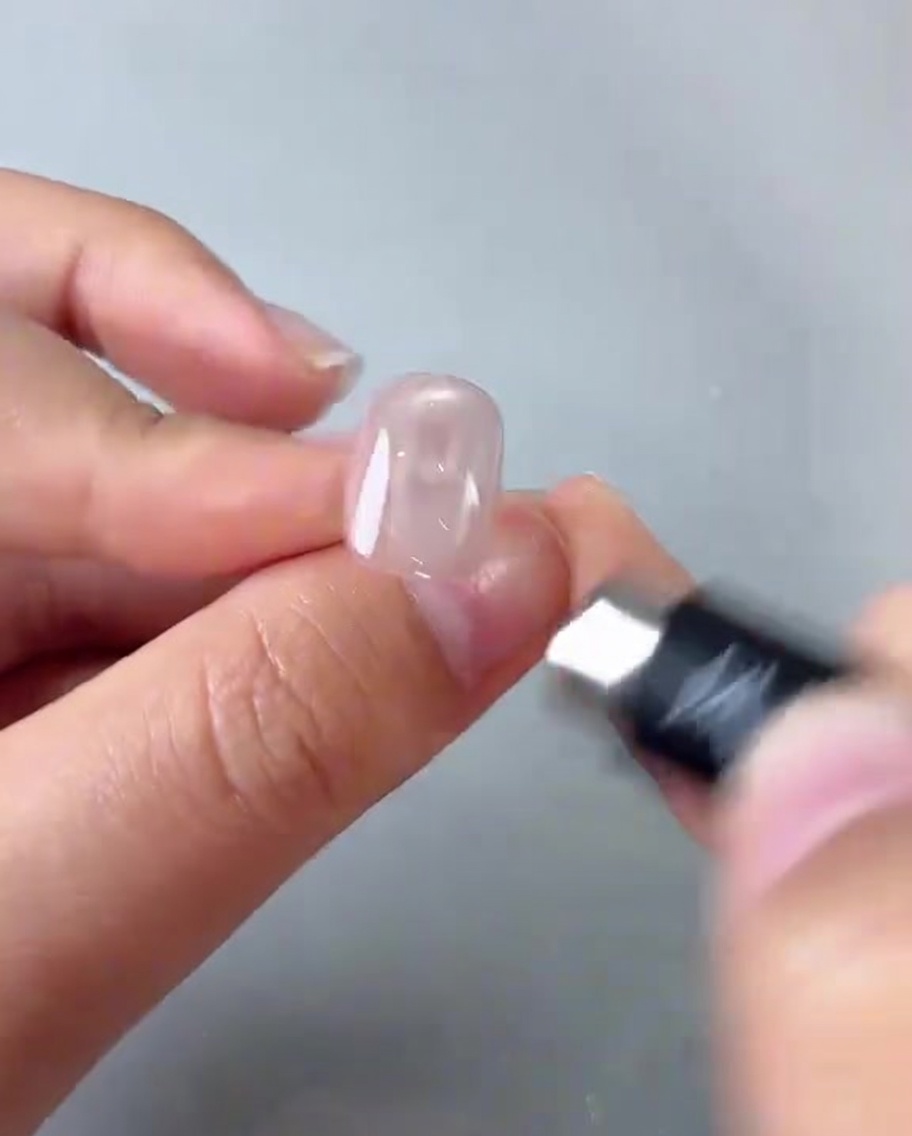 Nahaufnahme einer Hand, die einen kleinen zylindrischen Magneten über einen glänzenden rosa Nagel hält und eine helle, kreisförmige Reflexion erzeugt.