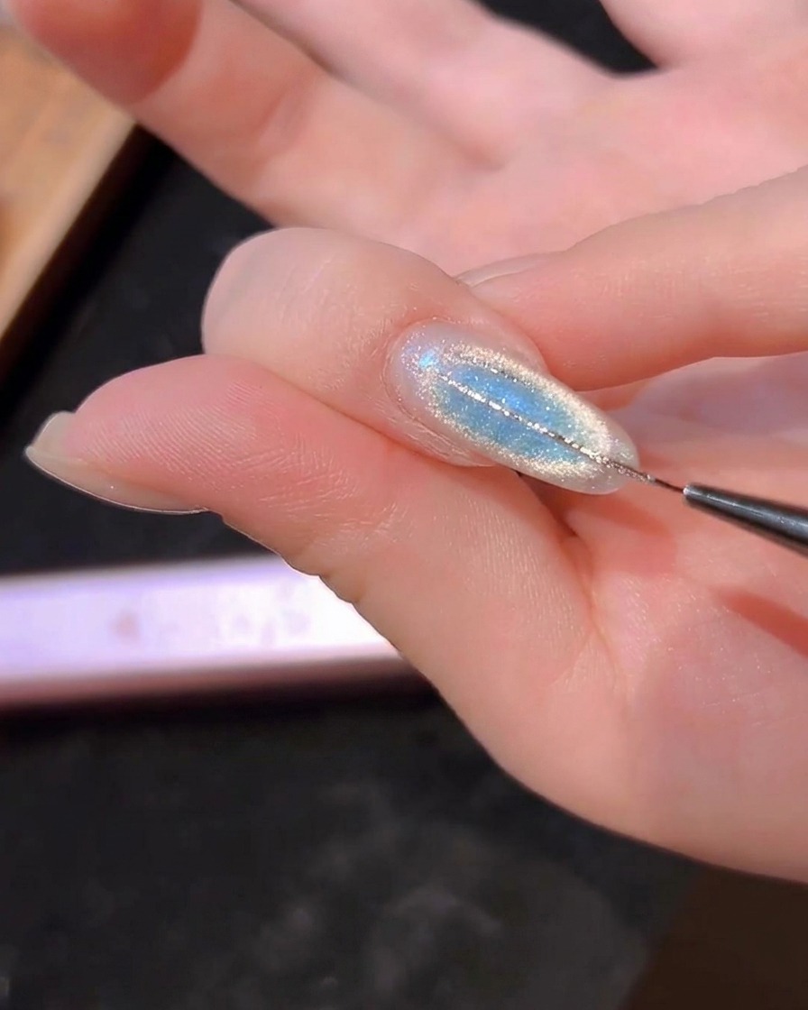 Ein feiner Liner-Pinsel zeichnet dünne silberne Linien auf einen blauen Cat-Eye-Nagel.