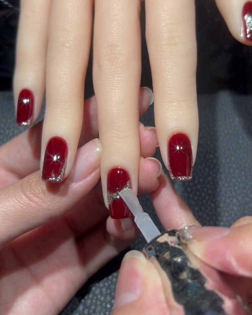 Ein Pinsel trägt klaren Top Coat auf einen roten Nagel mit Glitter-Design auf, wobei der zentrale Strassstein umgangen wird.