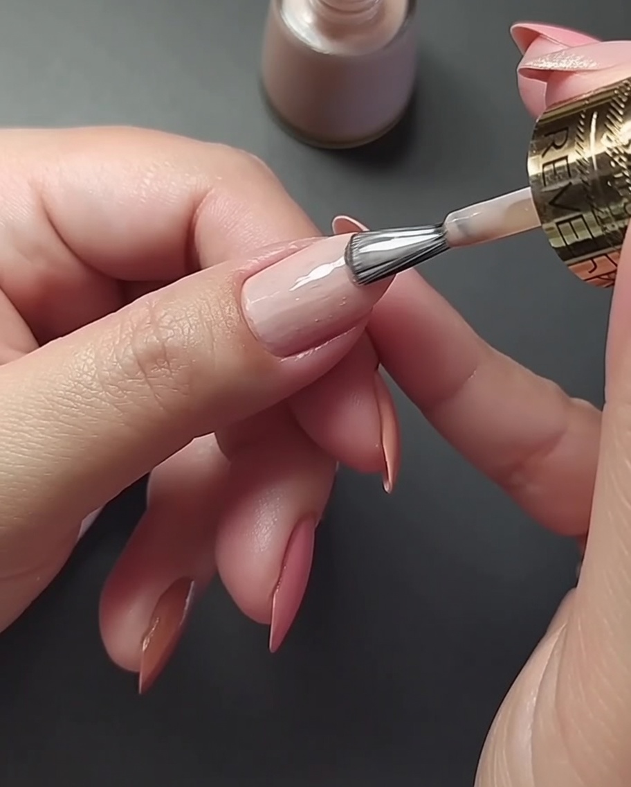 Nahaufnahme, wie eine dünne metallische Goldlinie vertikal über die Mitte eines hellen Nude-Daumennagels gemalt wird.