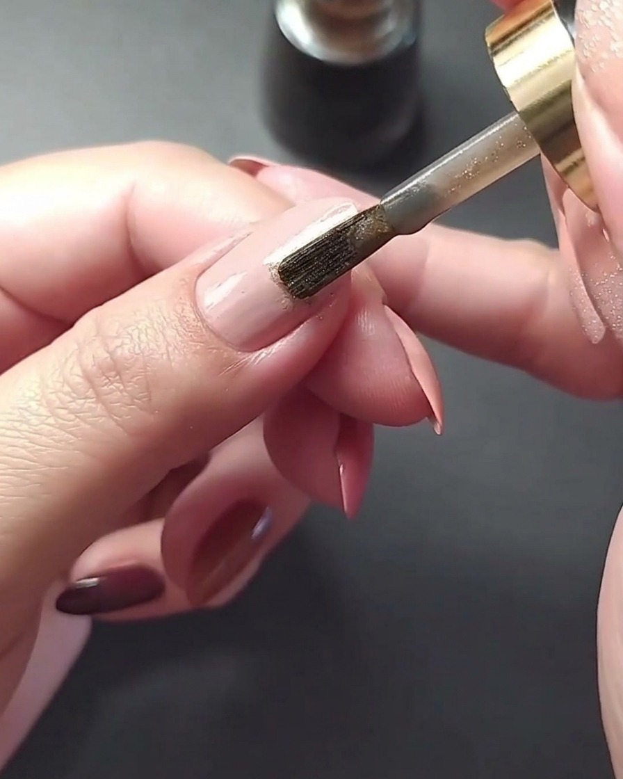 Makroaufnahme, wie eine dünne metallische Goldlinie vertikal mittig auf einen mauvefarbenen Fingernagel gemalt wird.