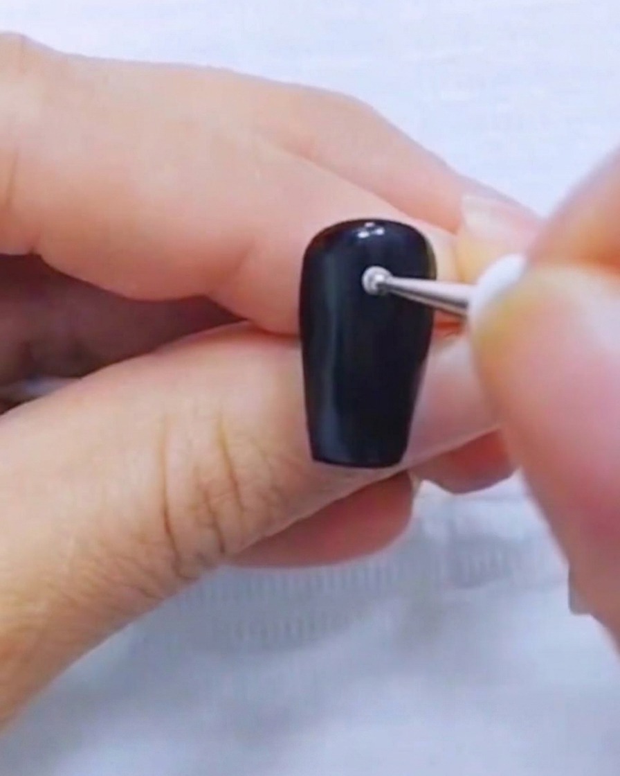 Nahaufnahme eines Dotting-Tools, das einen kleinen weißen Punkt auf einen schwarzen Nagel setzt.