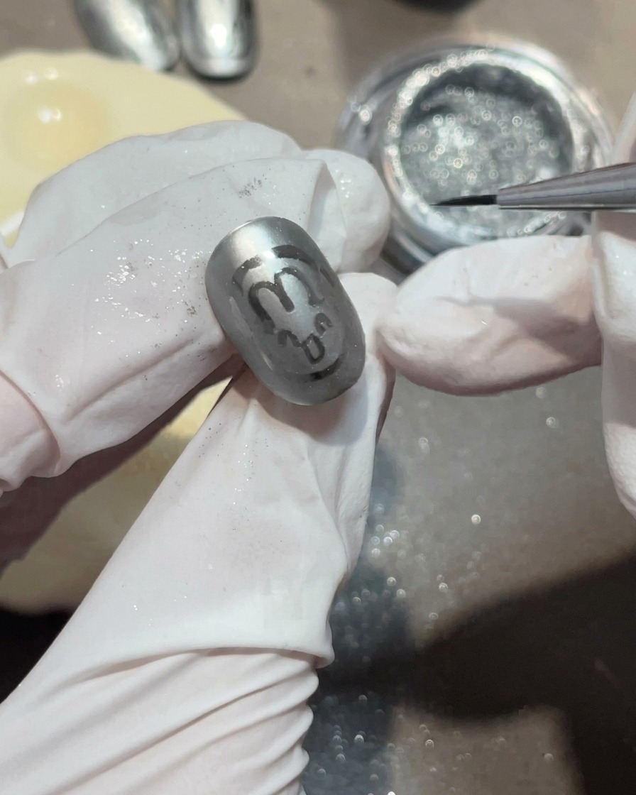Nahaufnahme eines Liner-Pinsels, der einen kleinen, klaren Gel-Tropfen auf einen matten grauen Nagel aufträgt, um einen Wassertropfen zu bilden.