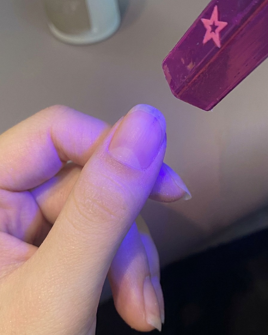 Eine Hand platziert einen Fingernagel unter einer kleinen, violetten UV-Handlampe, um den Base Coat auszuhärten.