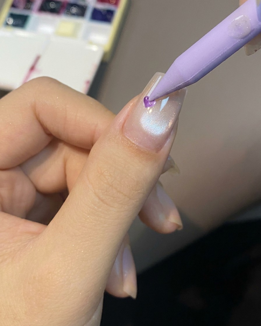 Ein spitzes Werkzeug platziert einen kleinen Tropfen durchscheinendes Gel in die Mitte eines vorbereiteten Cat-Eye-Nagels.