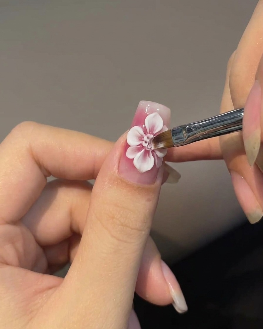 Ein abgeschrägter Nail-Art-Pinsel malt verblasste weiße äußere Blütenblätter um eine rote Rosenmitte auf einem rosa Nagel.