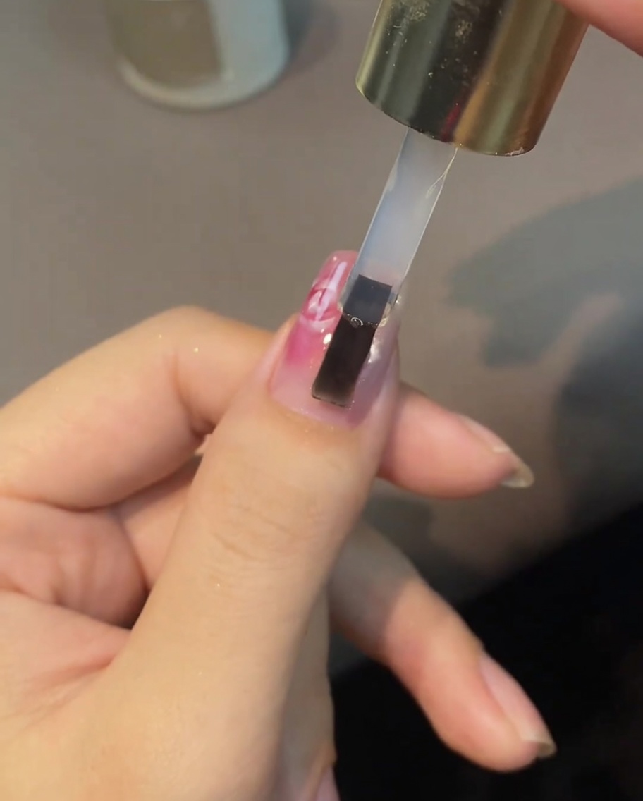 Ein Pinsel trägt einen klaren Top Coat über ein gemaltes weißes und rosa Rosennageldesign auf.