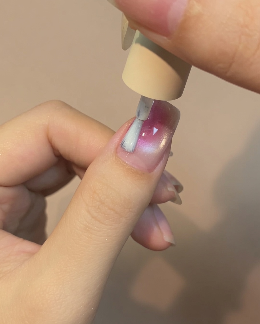 Ein Pinsel trägt eine dünne Schicht klaren Top Coat über ein rosa-weißes Rosen-Cat-Eye-Nageldesign auf.