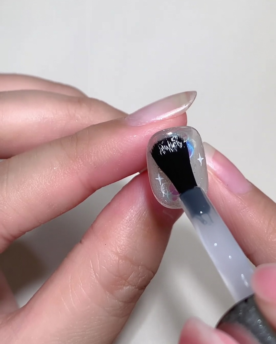 Ein Nagelpinsel trägt klares Builder-Gel über einem dekorierten Nagel auf, um die Oberfläche zu ebnen.