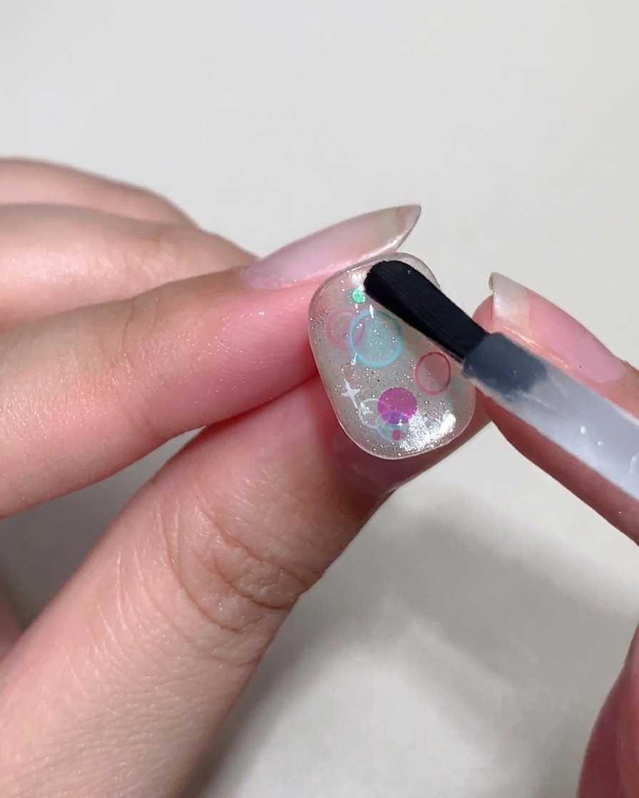 Ein Detailpinsel verwendet dicken Top Coat, um kleine, erhabene Blasen über den Pailletten auf dem Nagel zu formen.
