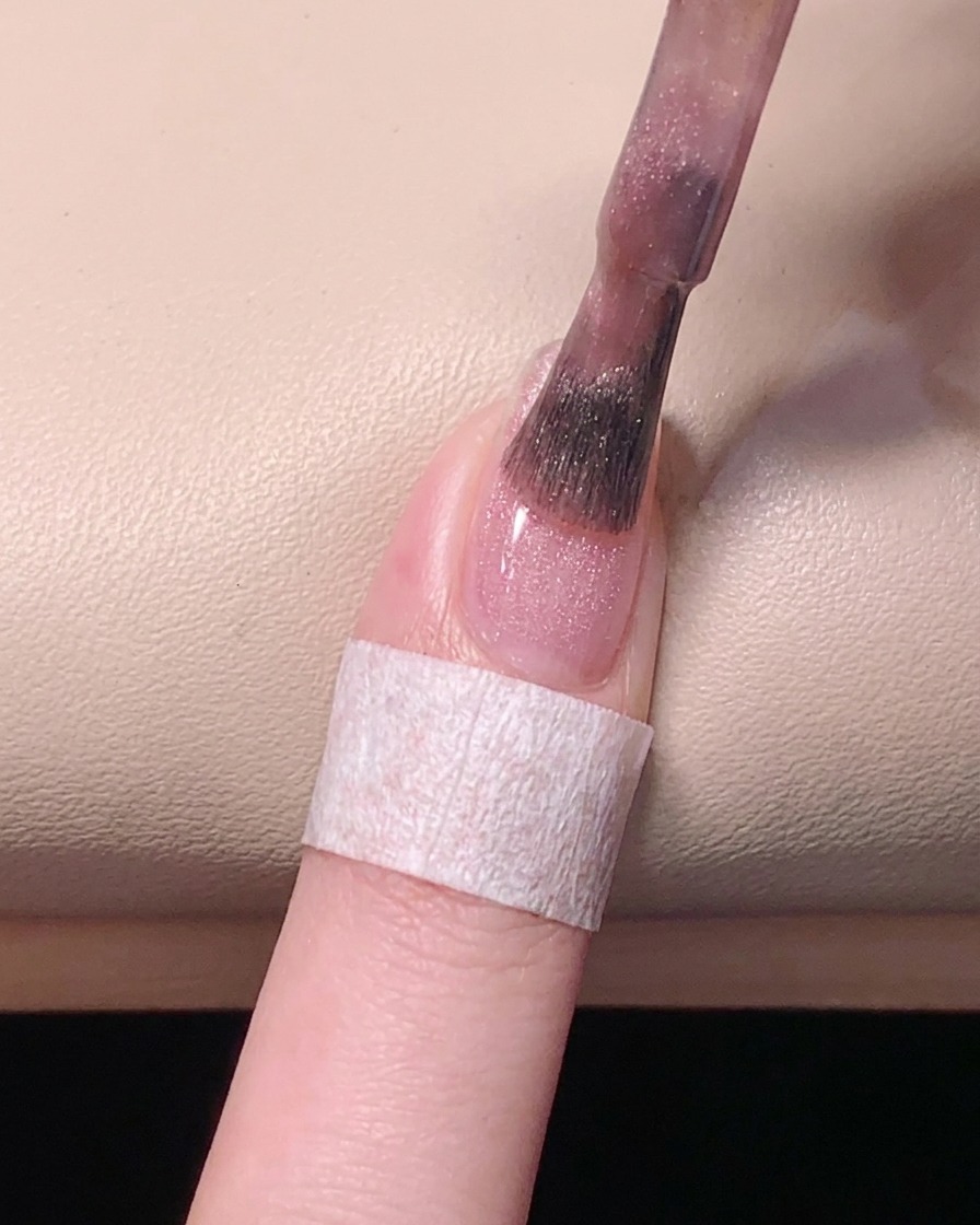 Ansicht von oben, wie ein Pinsel den pinken Gel-Lack auf der Nagelplatte für ein ebenmäßiges Finish glatt streicht.