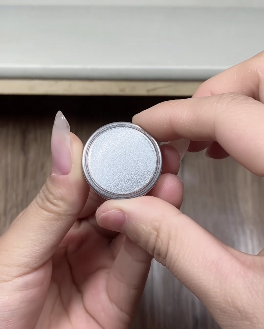 Hände halten einen kleinen, runden Behälter mit silbernem Chrom-Puder, der gerade geöffnet wurde.