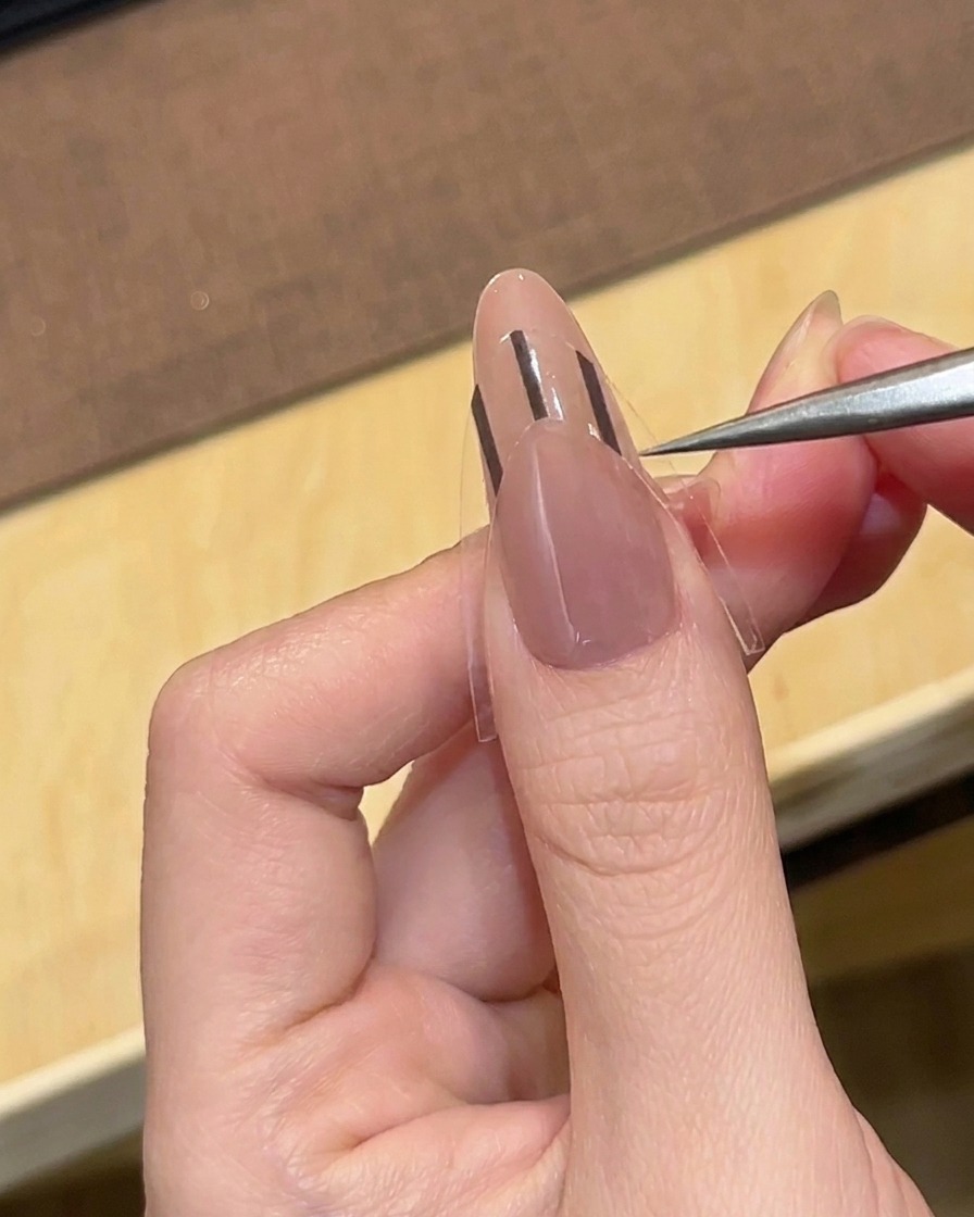 Eine Pinzette platziert einen schwarzen French-Tip-Aufkleber auf einem nackten, mandelförmigen Nagel.
