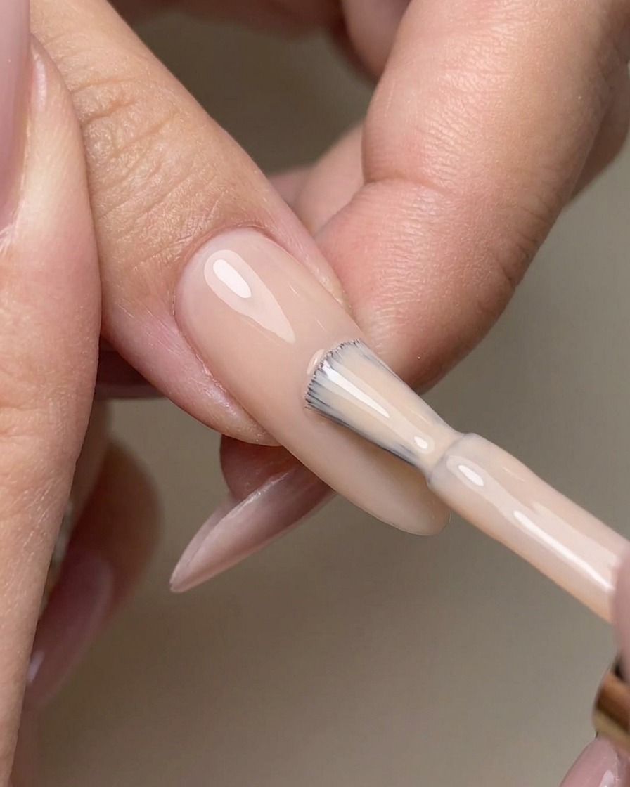 Ein Pinsel trägt einen klaren No-Wipe Top Coat gleichmäßig auf einen lackierten Nude-Gelnagel auf.