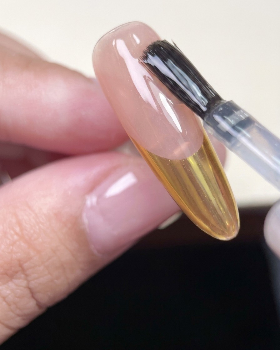 Ein Nagelpinsel trägt einen klaren, glänzenden Top Coat auf einen mandelförmigen Nagel mit goldener Chrome-French-Tip auf.