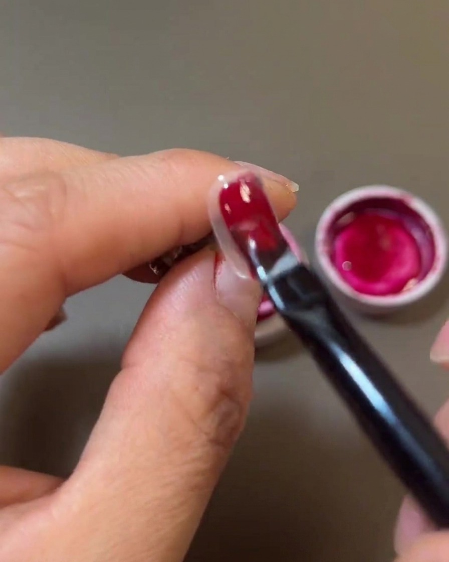 Nahaufnahme eines Nagelpinsels, der sattes, tiefrotes Gel auf die Nageloberfläche aufträgt.