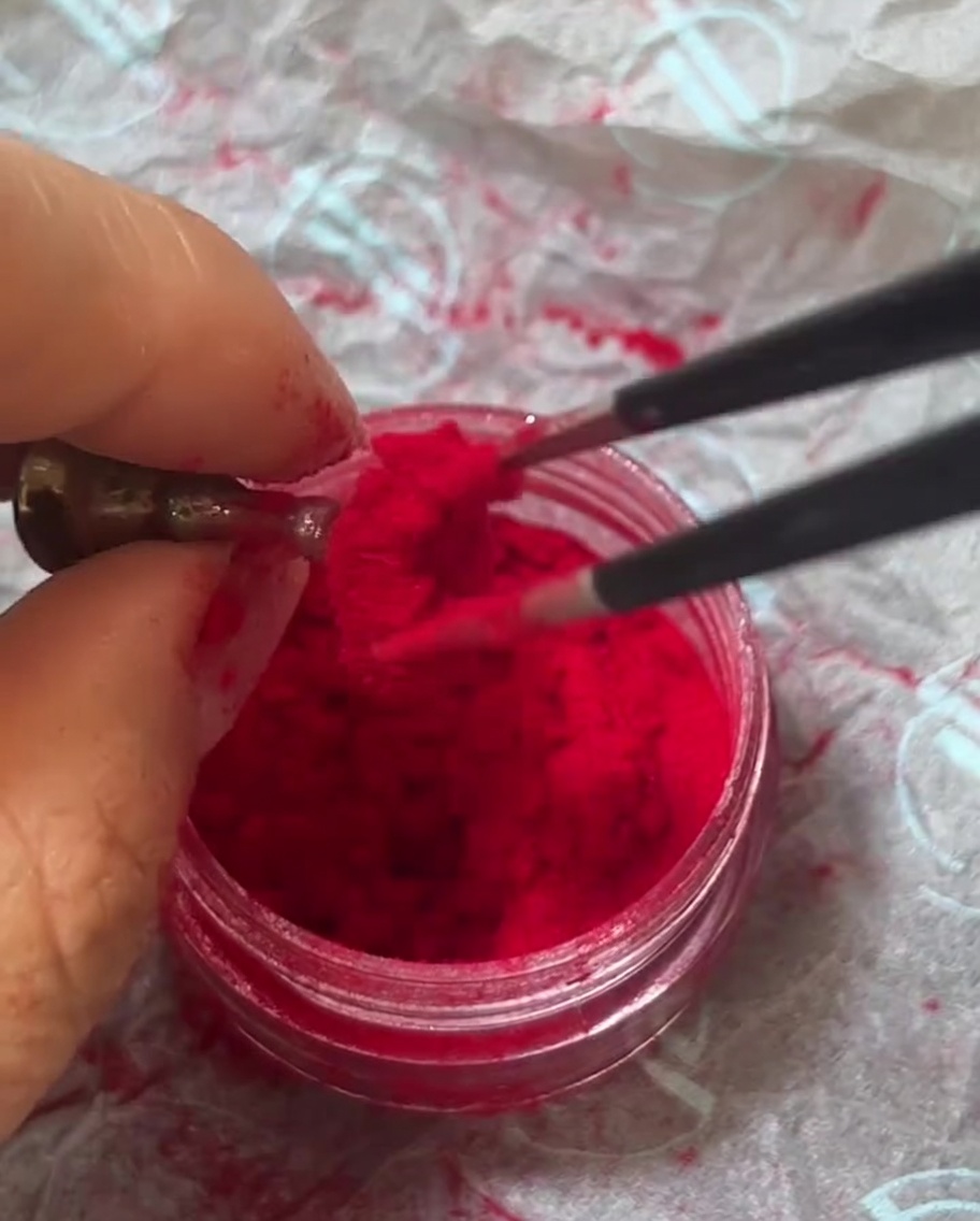 Eine Pinzette hält einen kleinen Büschel roter Samt-Flock-Fasern über ein Glas mit Pulver.