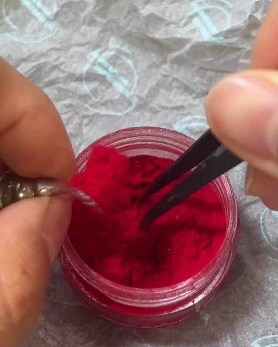 Die Pinzette streut vorsichtig rotes Plüsch-Pulver über die Oberfläche eines Nagels, der mit nassem Gel überzogen ist.