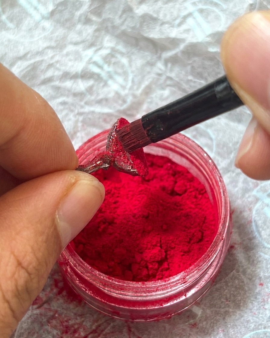 Ein Finger drückt vorsichtig auf das rote Plüsch-Pulver auf dem Nagel, um es im nassen Gel zu fixieren.