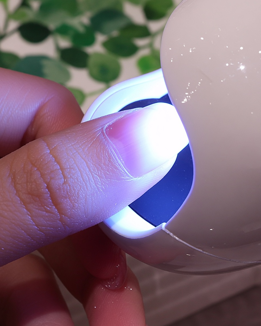 Ein Fingernagel mit einer strukturierten weißen Beschichtung unter dem violetten Licht einer kleinen UV/LED-Nagellampe.