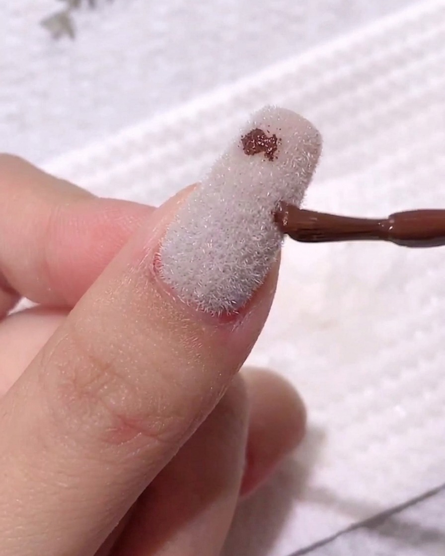 Ein Nagelpinsel platziert einen kleinen, dunkel kaffeefarbenen Geltropfen auf einer matten, weißen, samttexturierten Nageloberfläche.