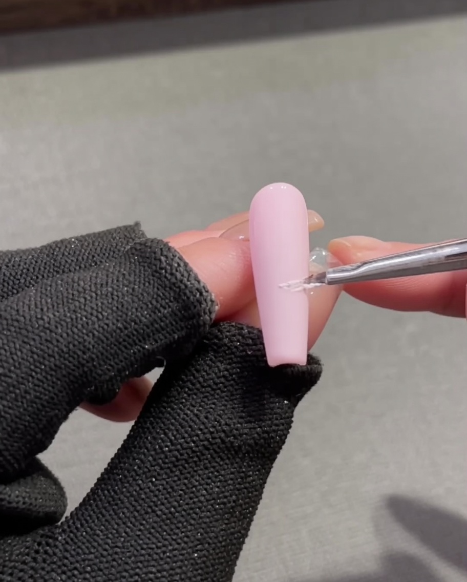 Ein feiner Detailpinsel trägt strukturiertes weißes Plaster-Gel auf, um eine French-Smile-Line auf dem rosa Nagel zu bilden.
