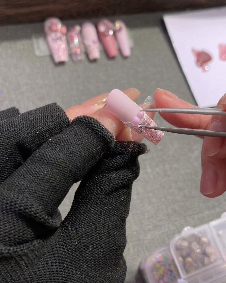Eine Pinzette positioniert kleine silberne Steinchen und eine pinkfarbene Schleife entlang der French-Linie.