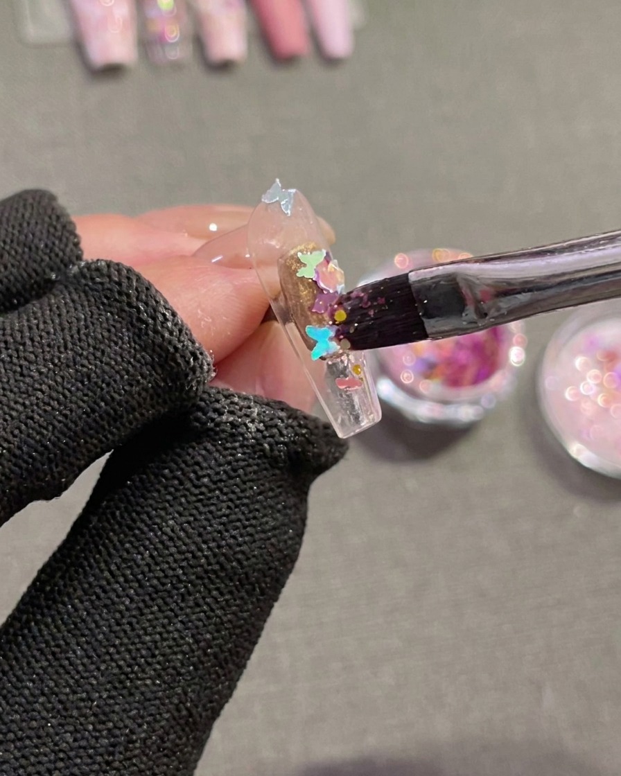 Ein Pinsel drückt schmetterlingsförmige holografische Pailletten in feuchtes Gel auf einem Nagel-Tip.