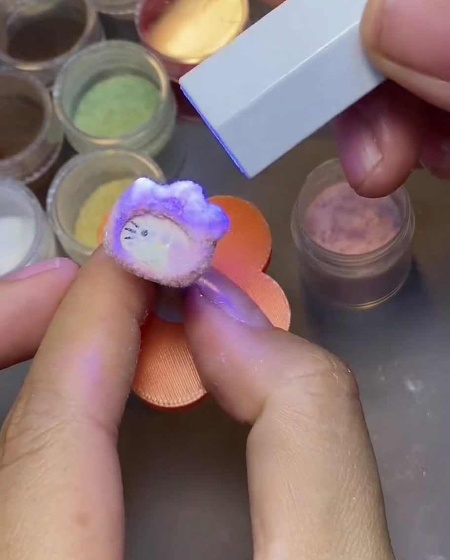 Eine Hand klopft an einen ausgehärteten Hello Kitty Nagelanhänger, um überschüssiges Samtpulver zu entfernen.