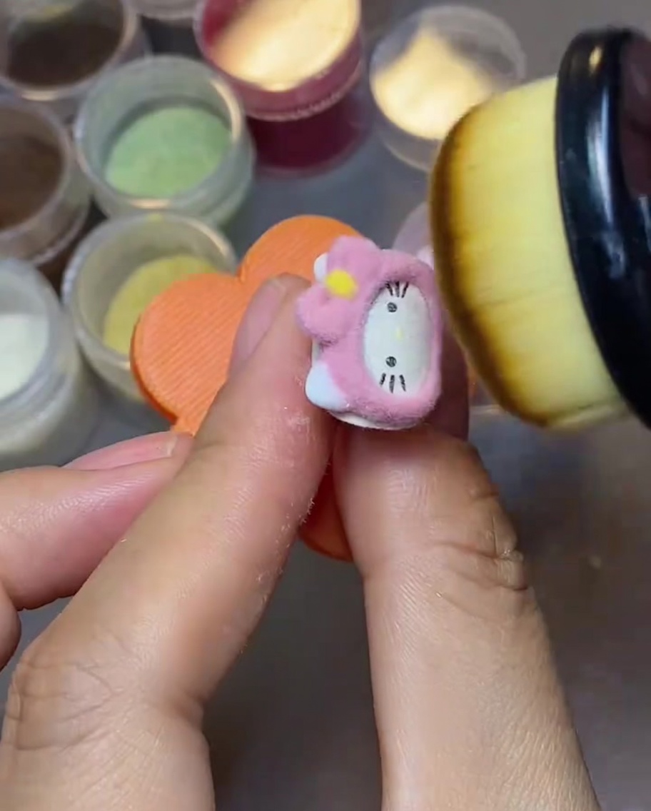 Eine Nahaufnahme eines flauschigen Make-up-Pinsels, der loses rosa Samtpulver von der Oberfläche eines Hello Kitty Nagelanhängers fegt.