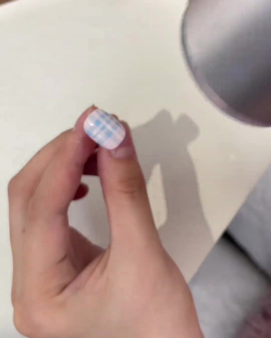 Klares Kristallpuder wird mit einem Metall-Nagelhautschieber über ein blaues Plaid-Muster auf einem Nagel gestreut.