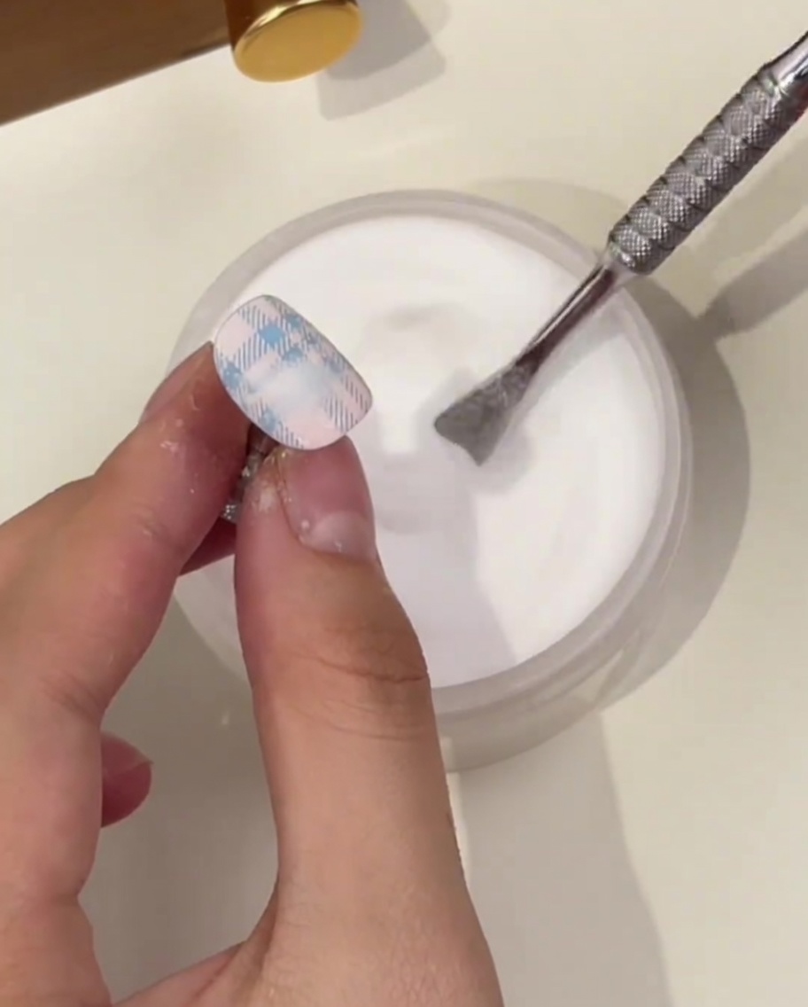 Nahaufnahme eines Nagels mit blauem Plaid-Muster, während Kristallpuder über dem Design gesättigt wird.