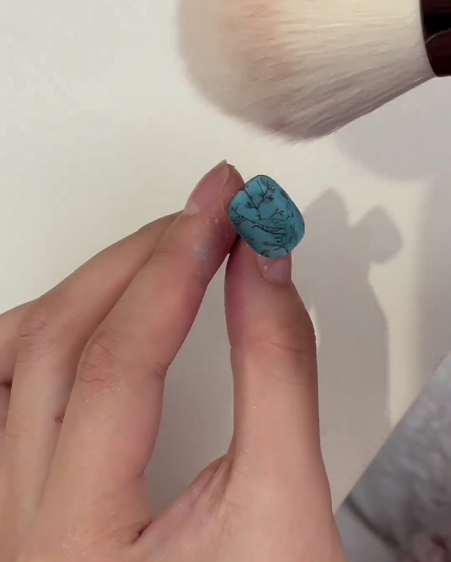 Ein flauschiger Pinsel staubt überschüssiges Puder von einem ausgehärteten türkisen Nagel ab.