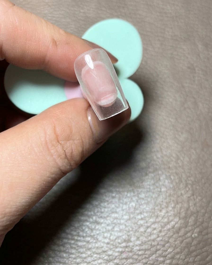 Nahaufnahme einer Hand, die einen klaren Nageltip hält und drei rosafarbene Gelpunkte als Grundlage zeigt.