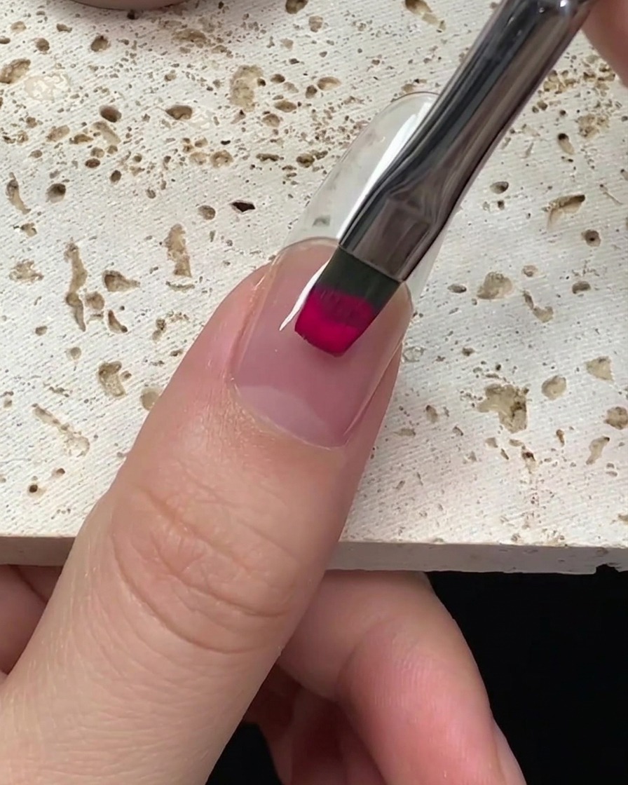 Nahaufnahme eines Pinsels, der vom Nagelmond aus leuchtend magentafarbenen Gellack auf einen Nagel aufträgt.