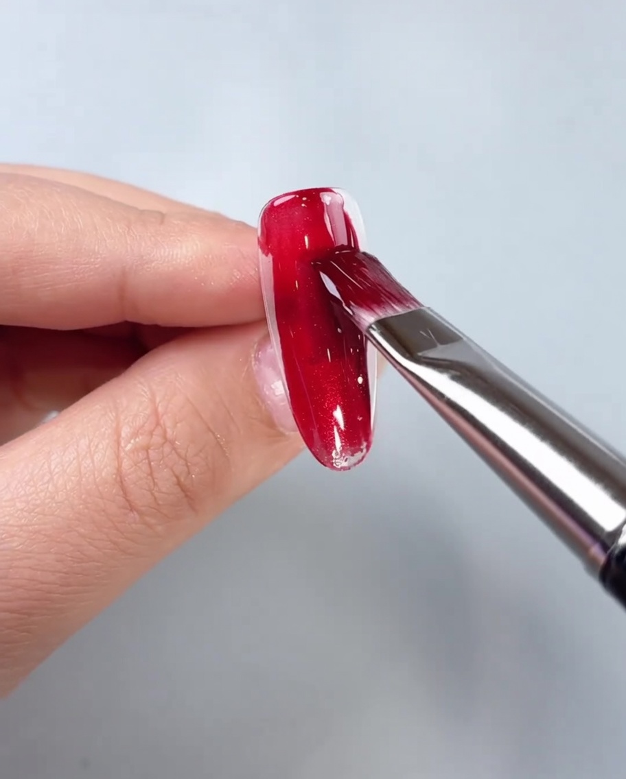 Ein Pinsel trägt rotes Cat-Eye-Gel auf die Mitte einer transparenten Nagelspitze auf.
