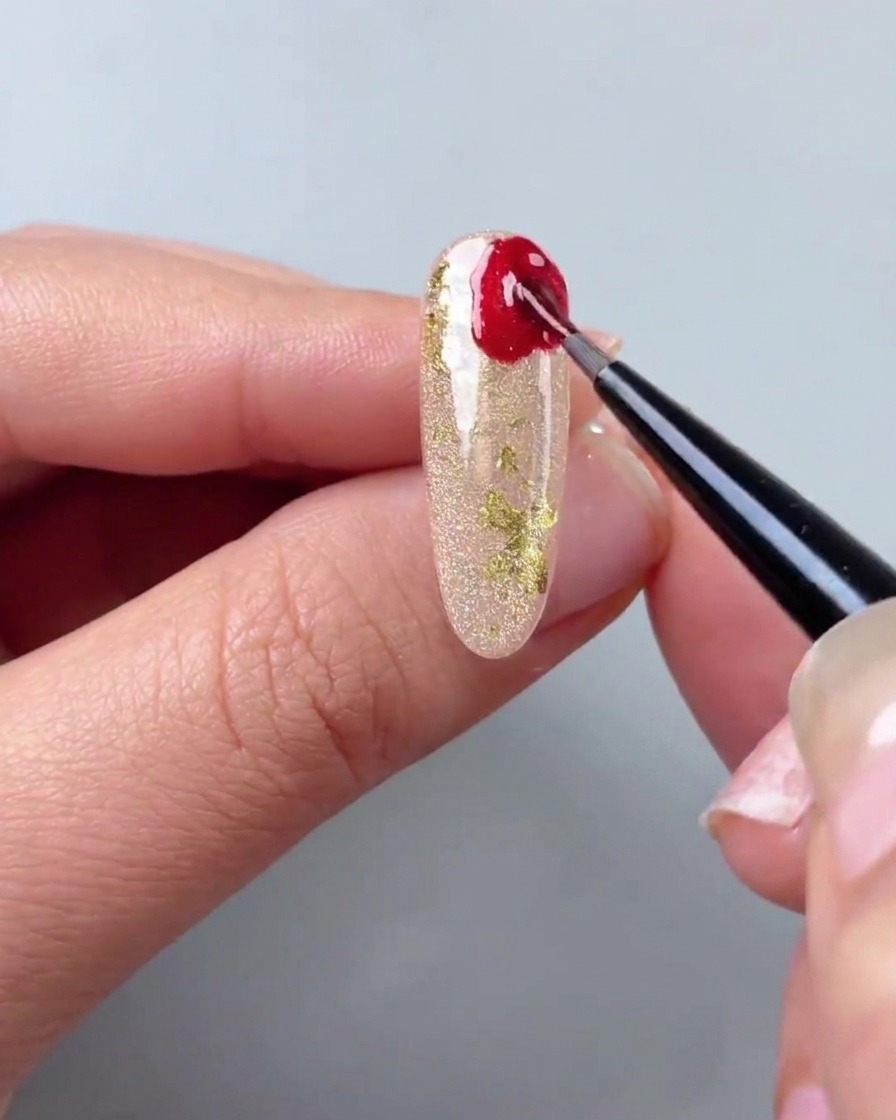 Ein feiner Detailpinsel trägt rotes Cat-Eye-Gel in unregelmäßigen Formen auf die Ecke eines schimmernden Goldnagels auf.