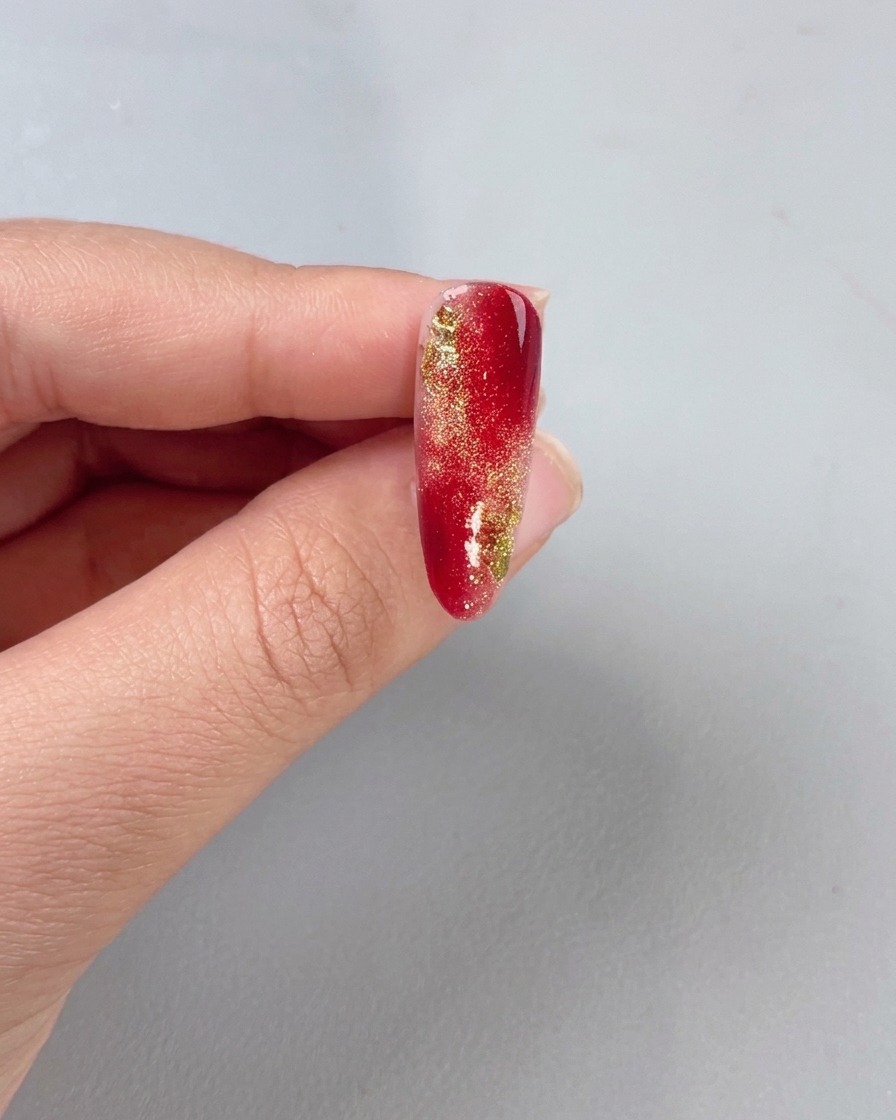 Ein Pinsel trägt einen klaren Top Coat über einen rot-goldenen Verlaufsnagel auf.