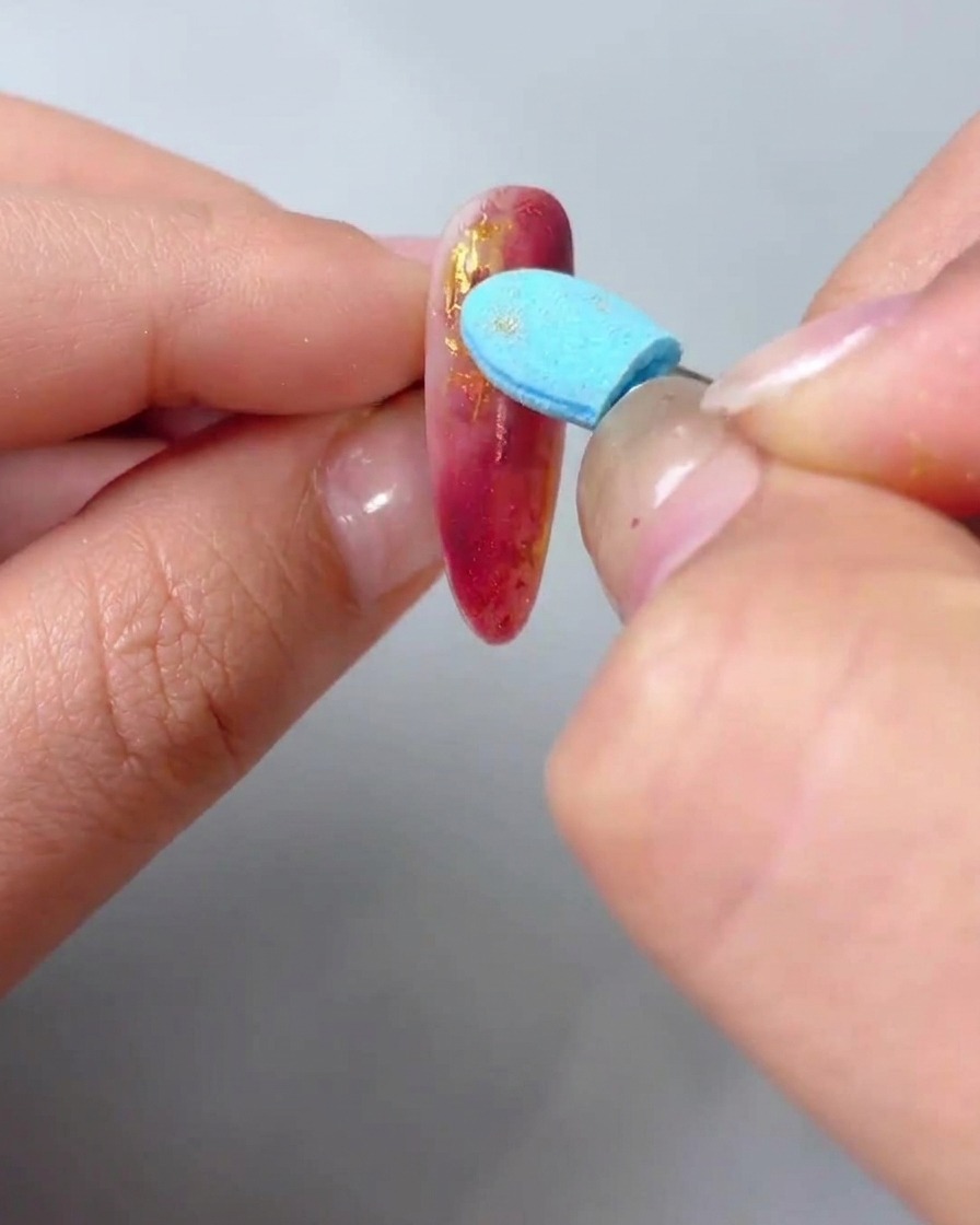 Nahaufnahme einer Hand, die mit einem blauen Schwamm-Applikator metallisches Goldpuder auf die erhabenen Texturlinien eines roten Marmornagels reibt.
