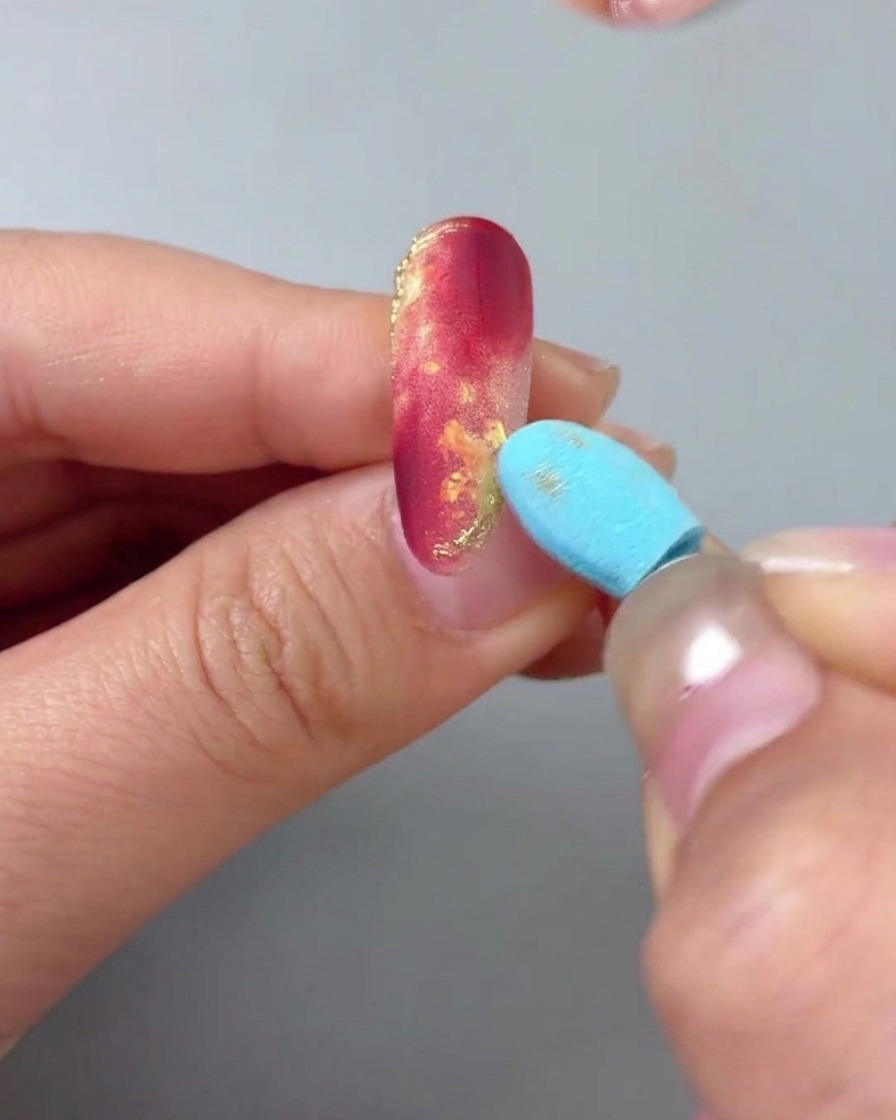 Nahaufnahme einer Hand mit einem rot-goldenen Marmornagel, während ein blaues Schwammwerkzeug sanft die Oberfläche abwischt, um überschüssiges Goldpuder zu entfernen.
