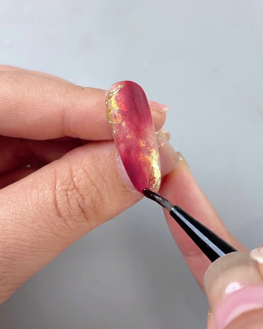Ein dünner Detailpinsel trägt einen klaren, isolierenden Top Coat über einen schön verzierten rot-goldenen Marmornagel auf.
