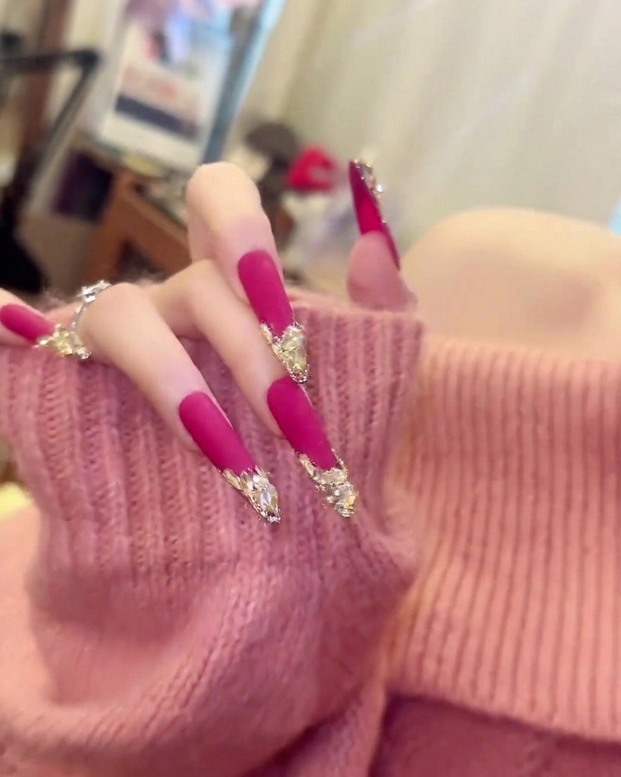 Eine Hand in einem hellrosa Pullover bewegt sich leicht, um die Länge und Krümmung langer, matter pinker Mandelnägel zu zeigen.