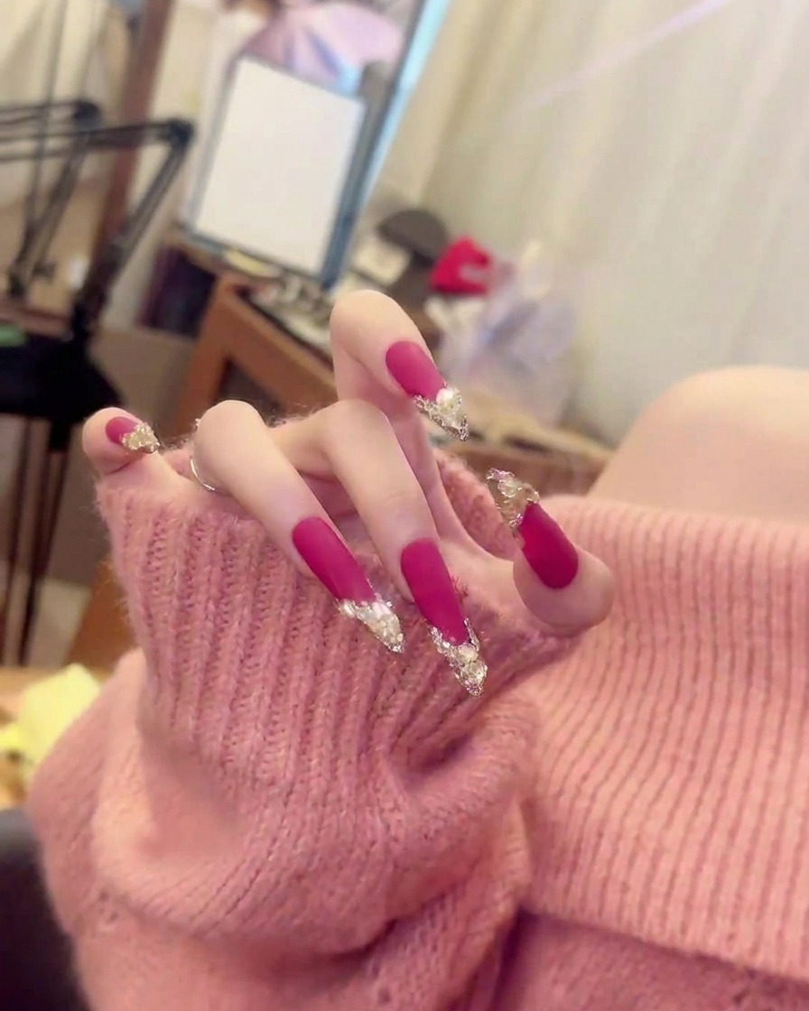 Eine scharfe Nahaufnahme eines einzelnen Fingers, die die präzise Platzierung von Kristall-Clustern auf mattem pinkem Lack zeigt.