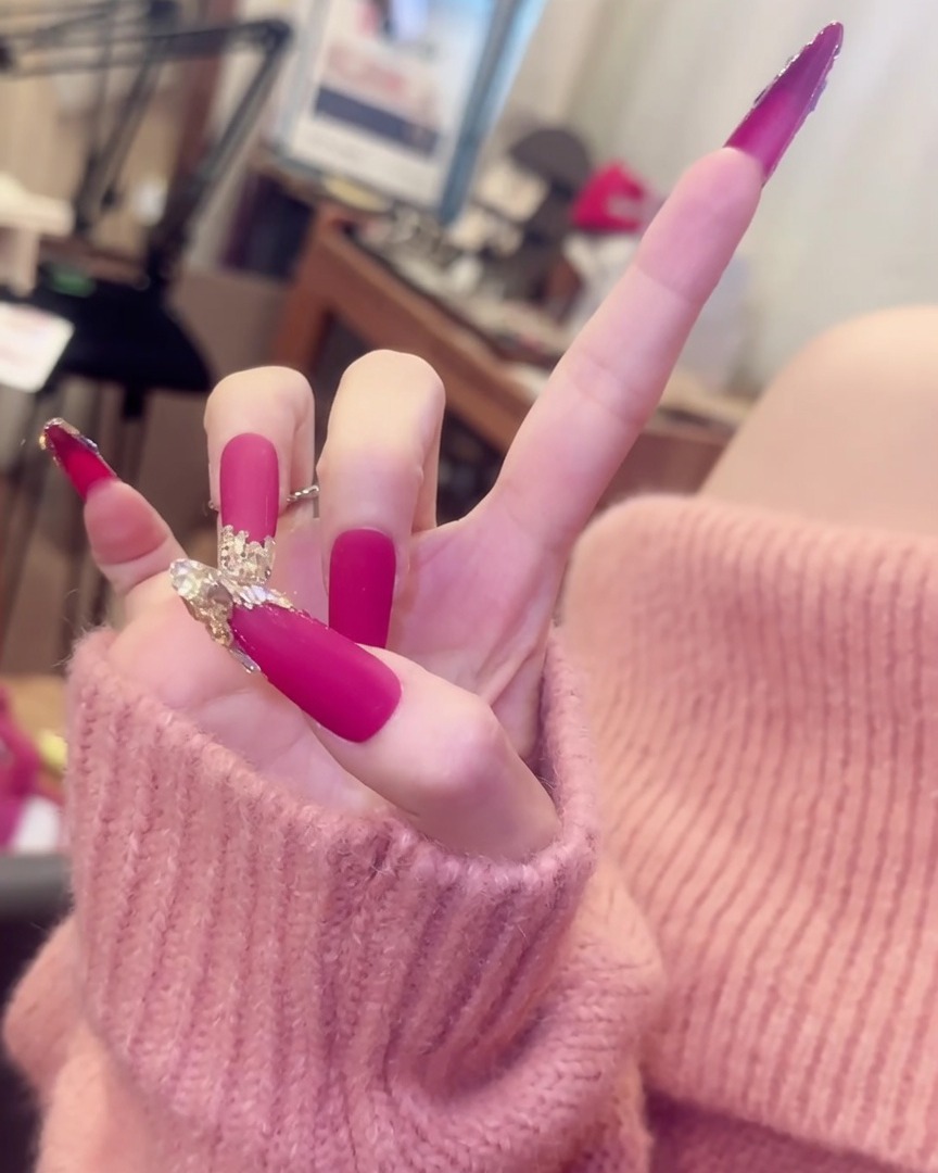 Eine Hand mit gespreizten Fingern, die lange matte pinke Nägel mit Kristallakzenten und eleganten Silberringen präsentiert.